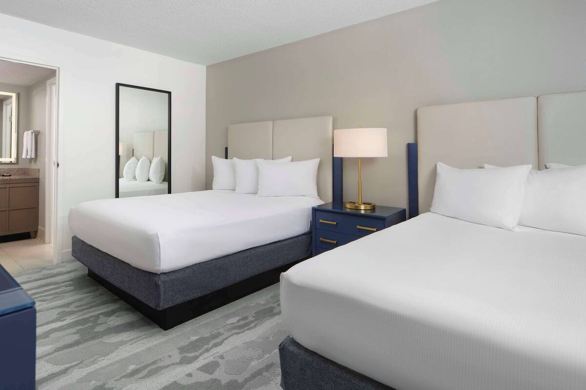 A hotel room with two beds, a nightstand with a lamp, a mirror, and a bathroom visible through an open door.