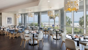 Un comedor moderno con mesas y sillas redondas, ventanas amplias con vistas a la playa y lámparas decorativas colgantes.