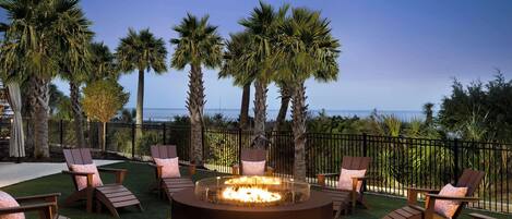 A circular fire pit surrounded by wooden chairs with cushions, set on artificial grass, with palm trees and a view of the ocean in the background.