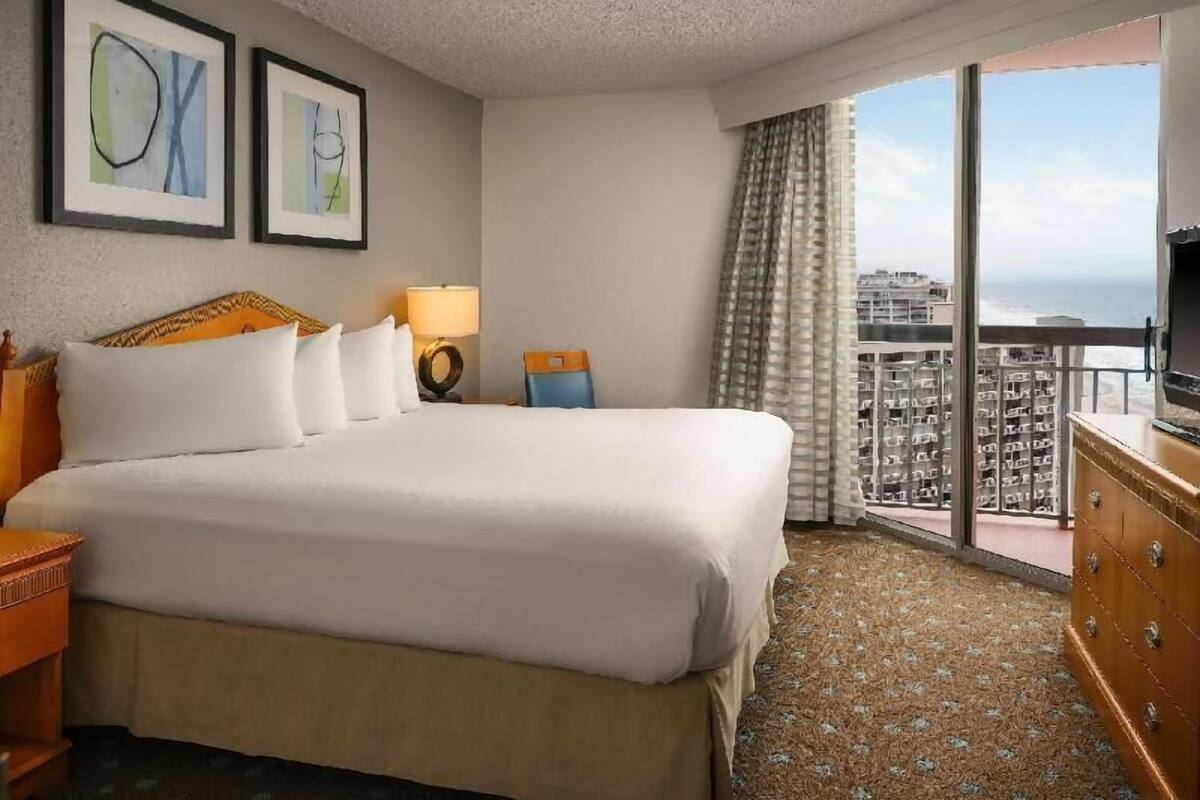A hotel room with a large bed, a dresser, a television, and a balcony with a city view.