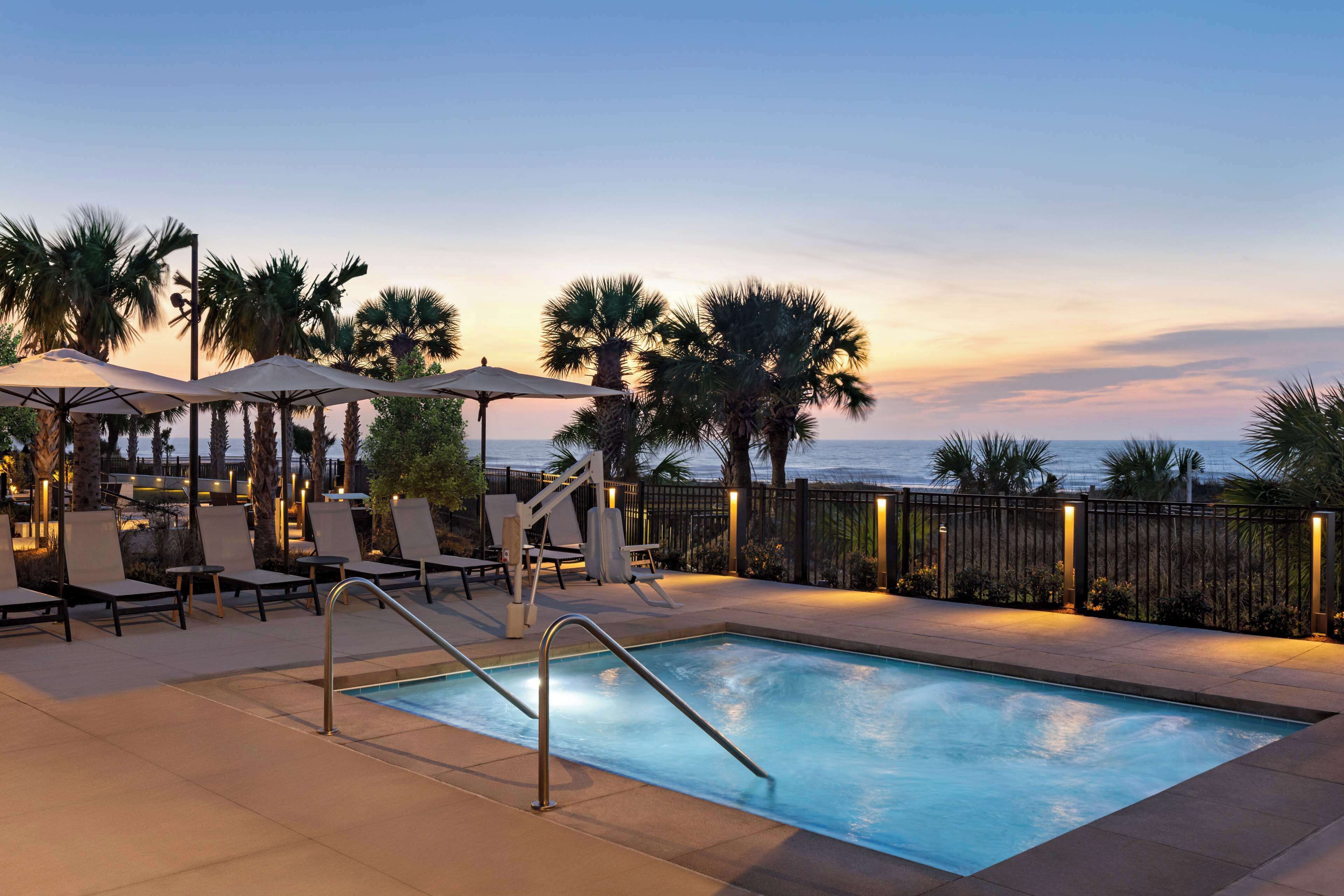 A pool area with lounge chairs and umbrellas, surrounded by palm trees and a view of the ocean.