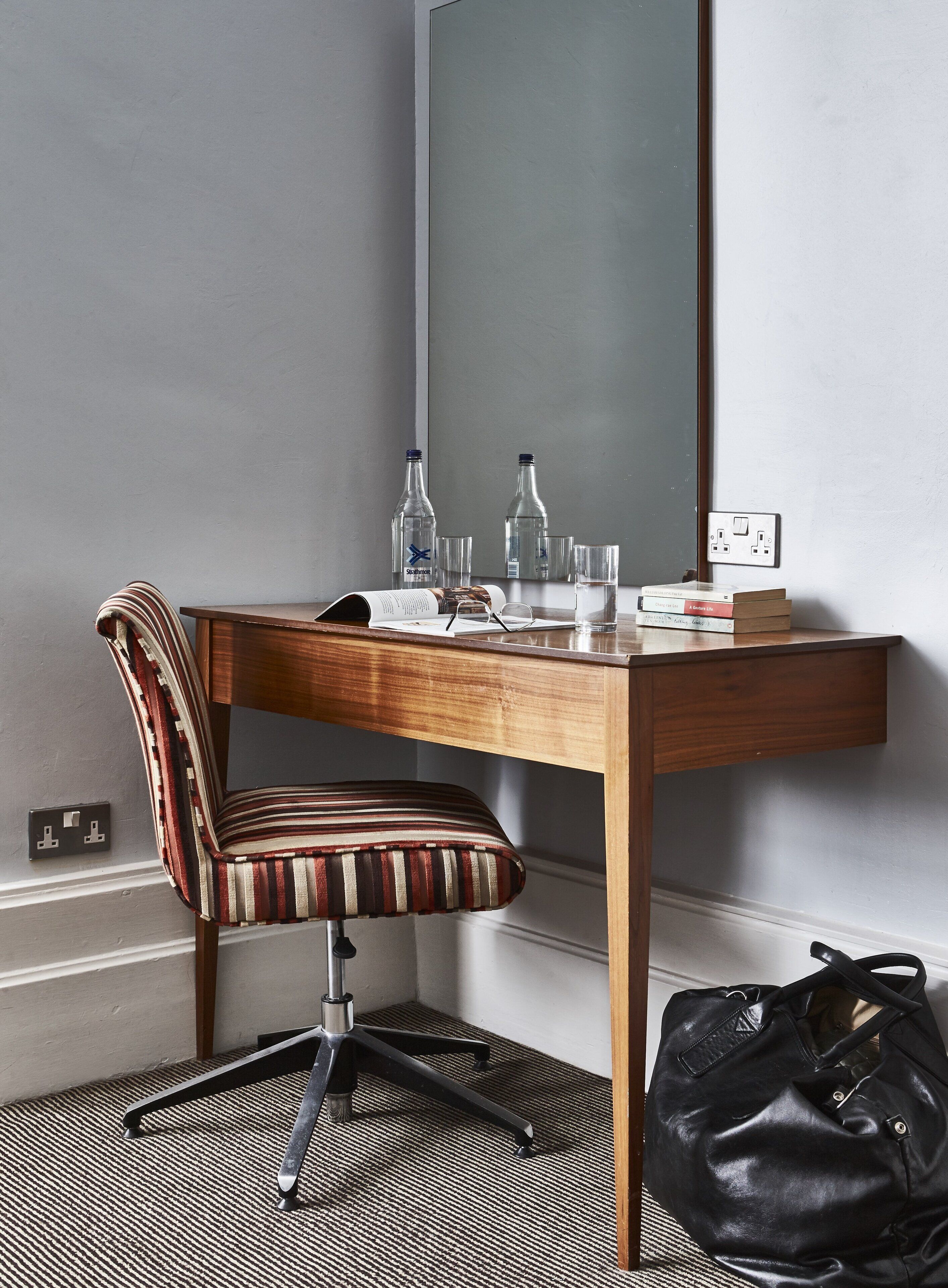 A hotel room with a wooden desk, striped chair, and a large mirror. There are two bottles, two glasses, and a few books on the desk. A black bag is on the floor next to the desk.
