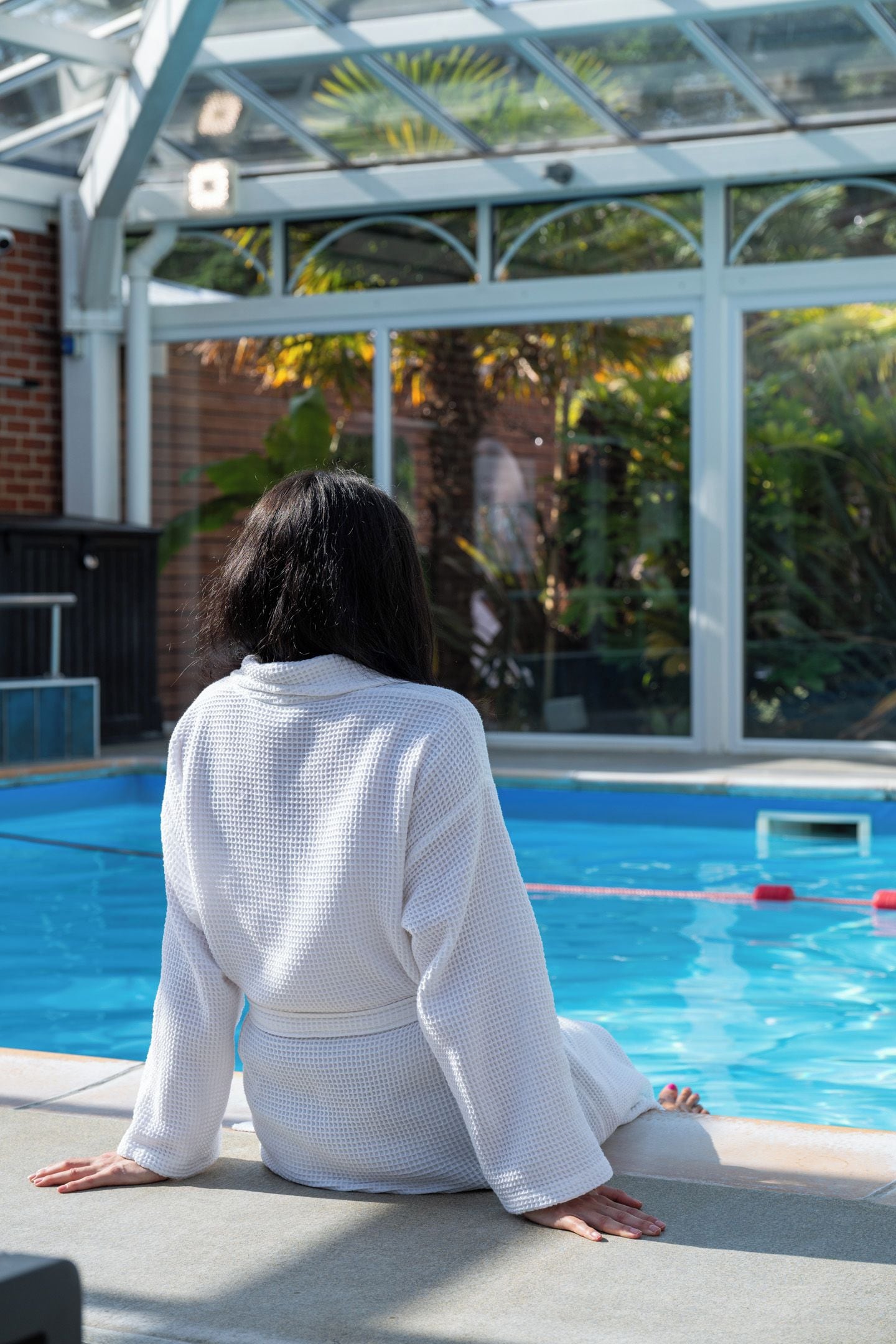En person sidder ved en pool under et glasoverdækket tag.