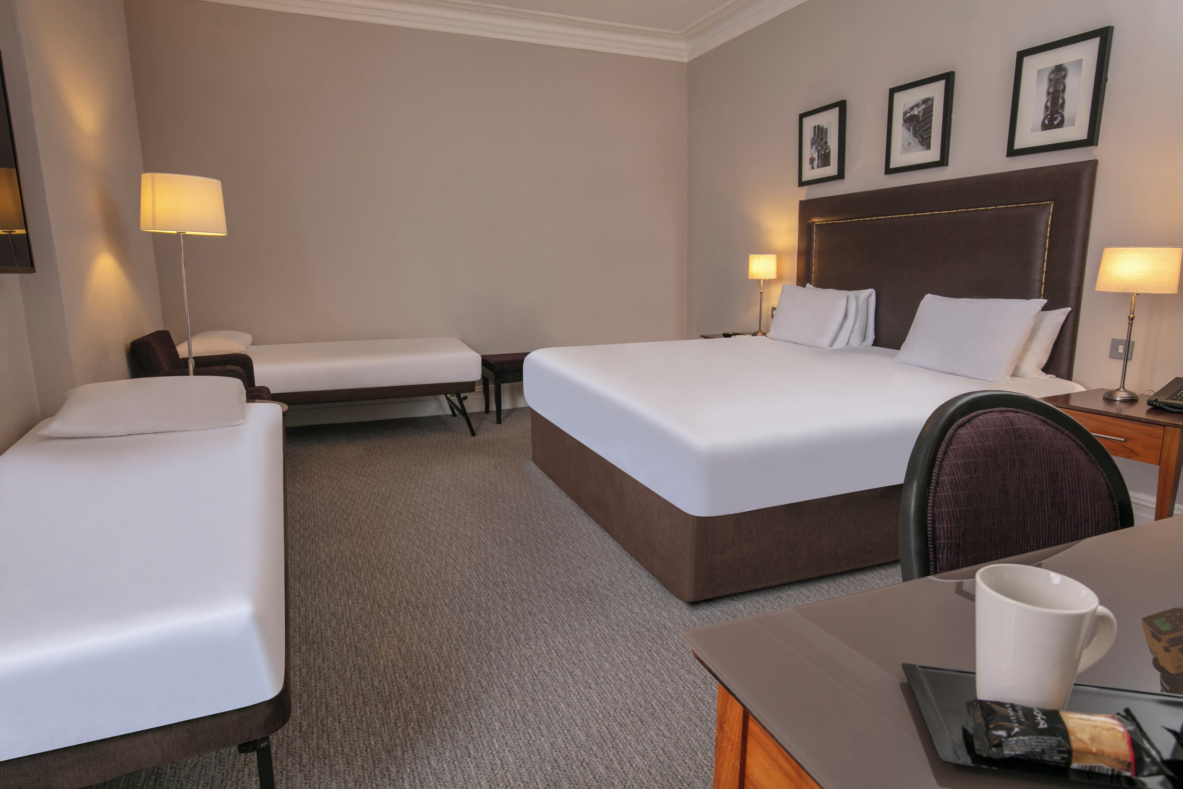 Une chambre d’hôtel avec deux lits simples, un lit double, un bureau avec une chaise et une table de chevet avec une lampe.