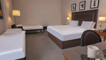 Une chambre d’hôtel avec deux lits simples, un lit double, un bureau avec une chaise et une table de chevet avec une lampe.
