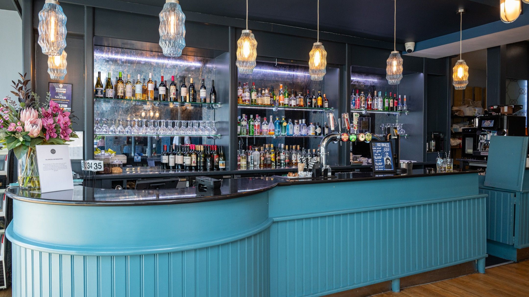Un espace bar avec un comptoir bleu, diverses bouteilles, des verres et des luminaires suspendus.