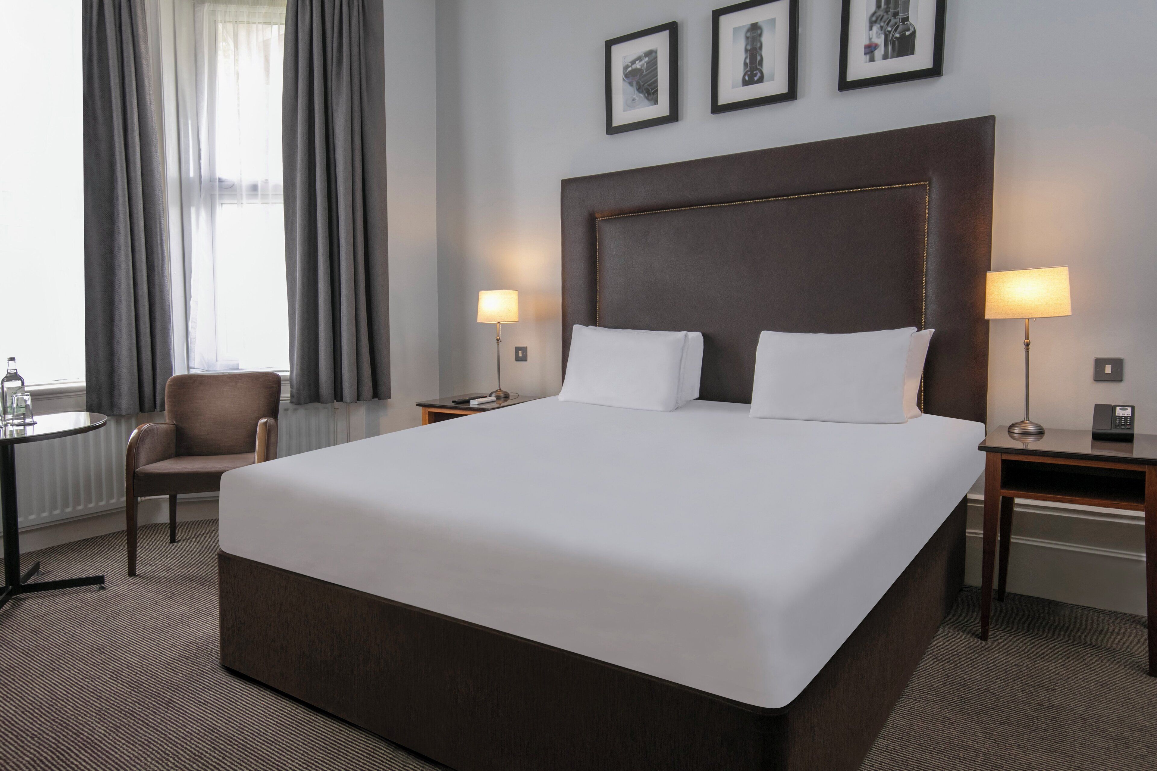 A hotel room with a large bed, two bedside tables with lamps, a chair, a small table, and a window with curtains.