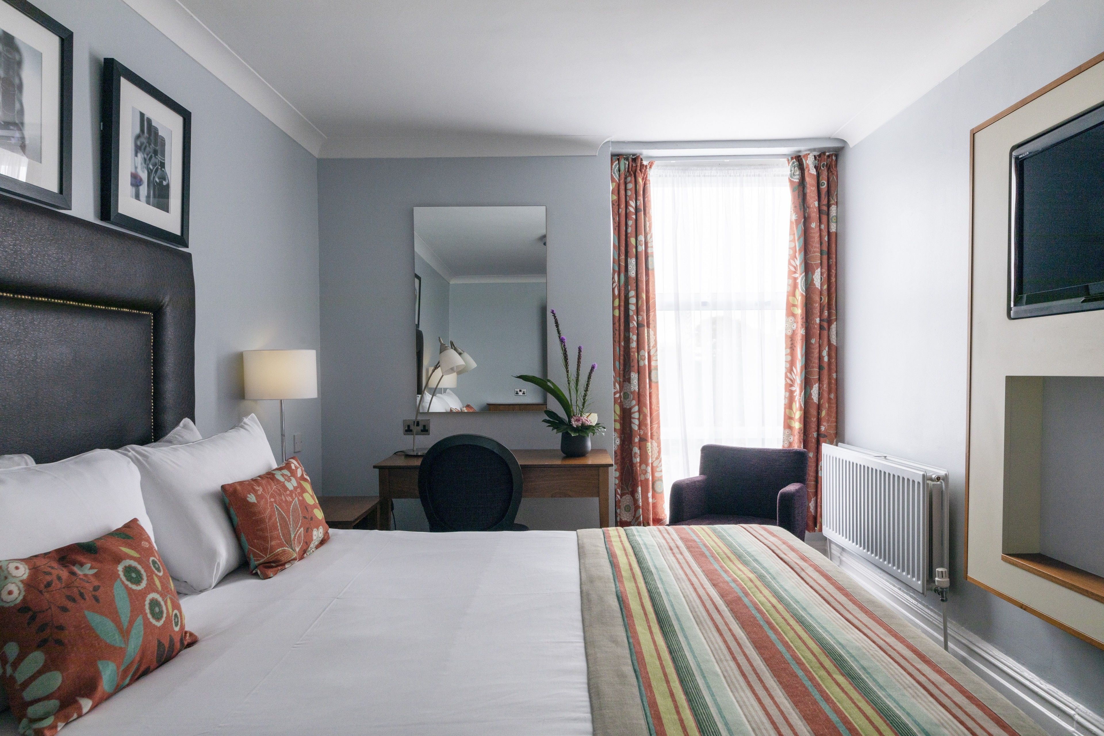 Habitación de hotel con cama, escritorio, silla, radiador y ventana con cortinas.