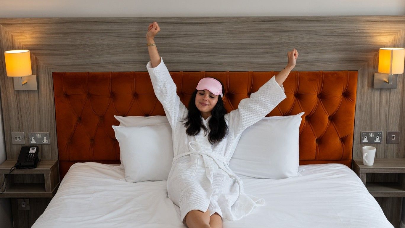 A hotel room with a tufted headboard, a bedside table with a lamp, a phone, and a cup.