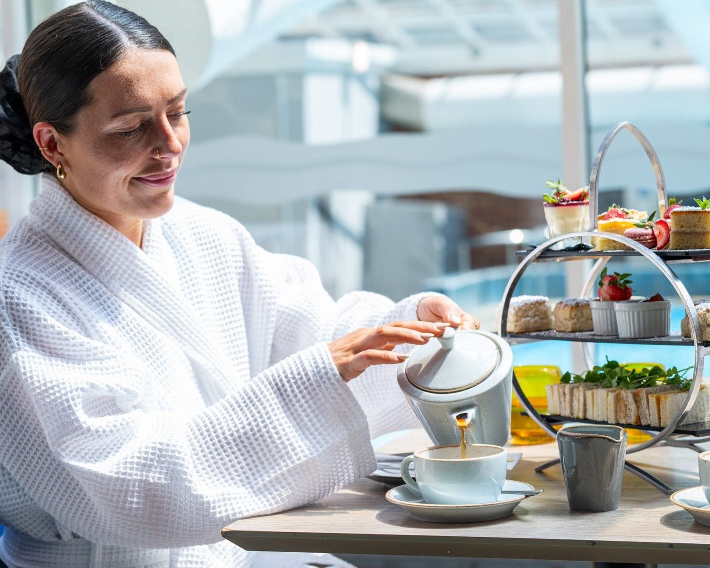 A person in a white robe pouring coffee into a cup, with a tiered dessert display in the background.