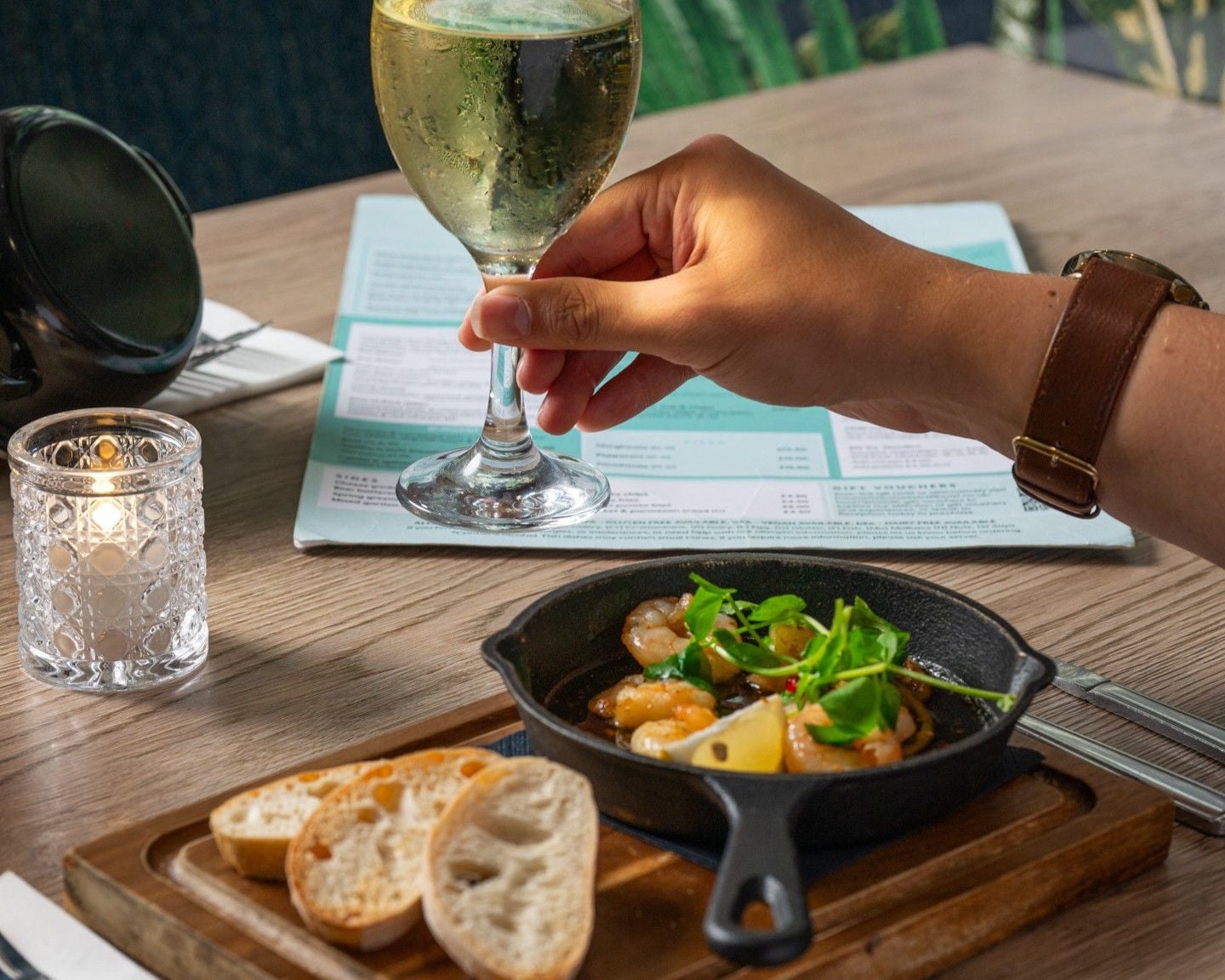 Une table à manger avec un menu, un verre de vin blanc, une poêle en fonte contenant des aliments et des tranches de pain.