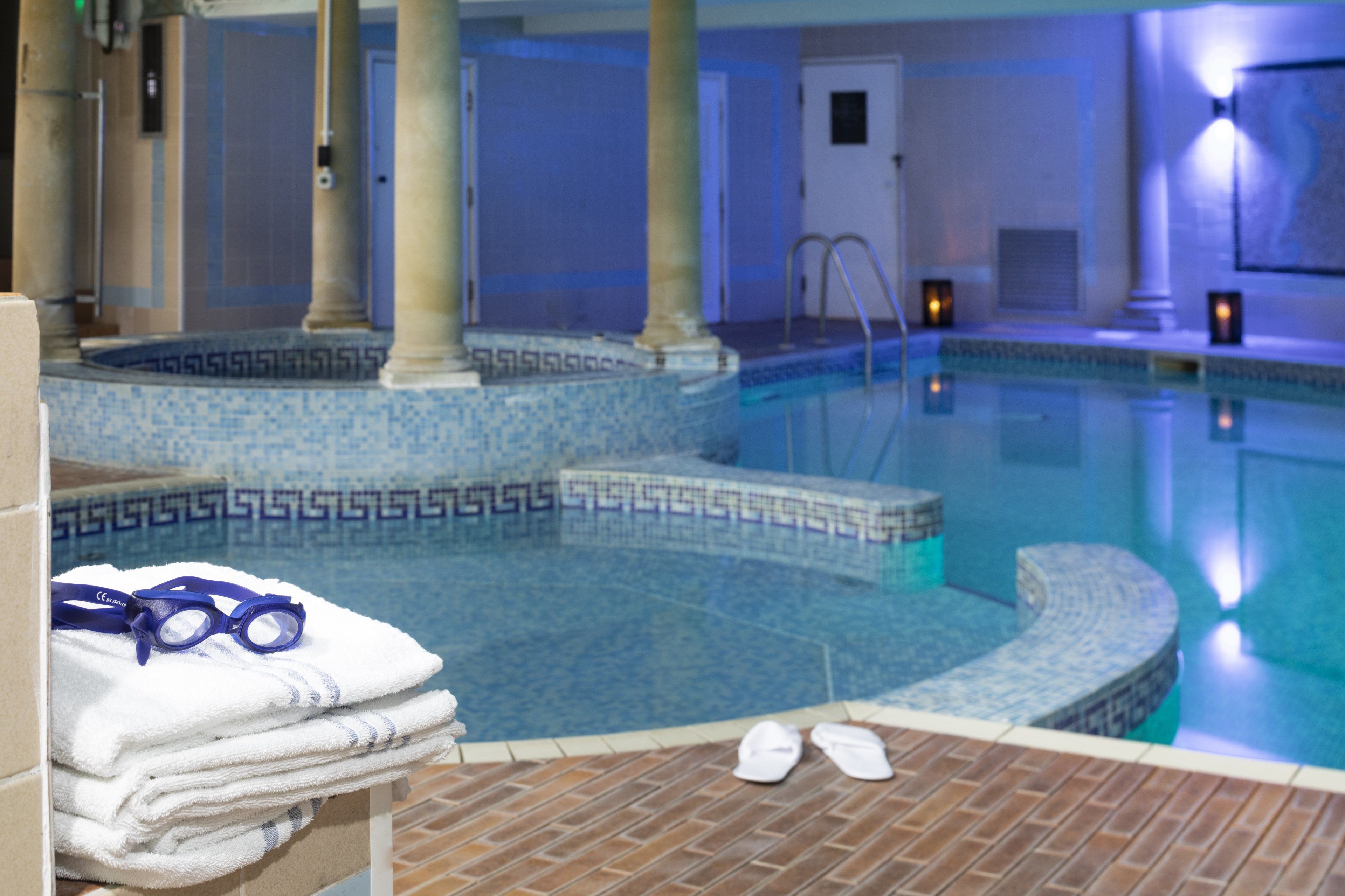 Una piscina interior con borde de azulejos, un jacuzzi pequeño y un juego de toallas y gafas a un lado.