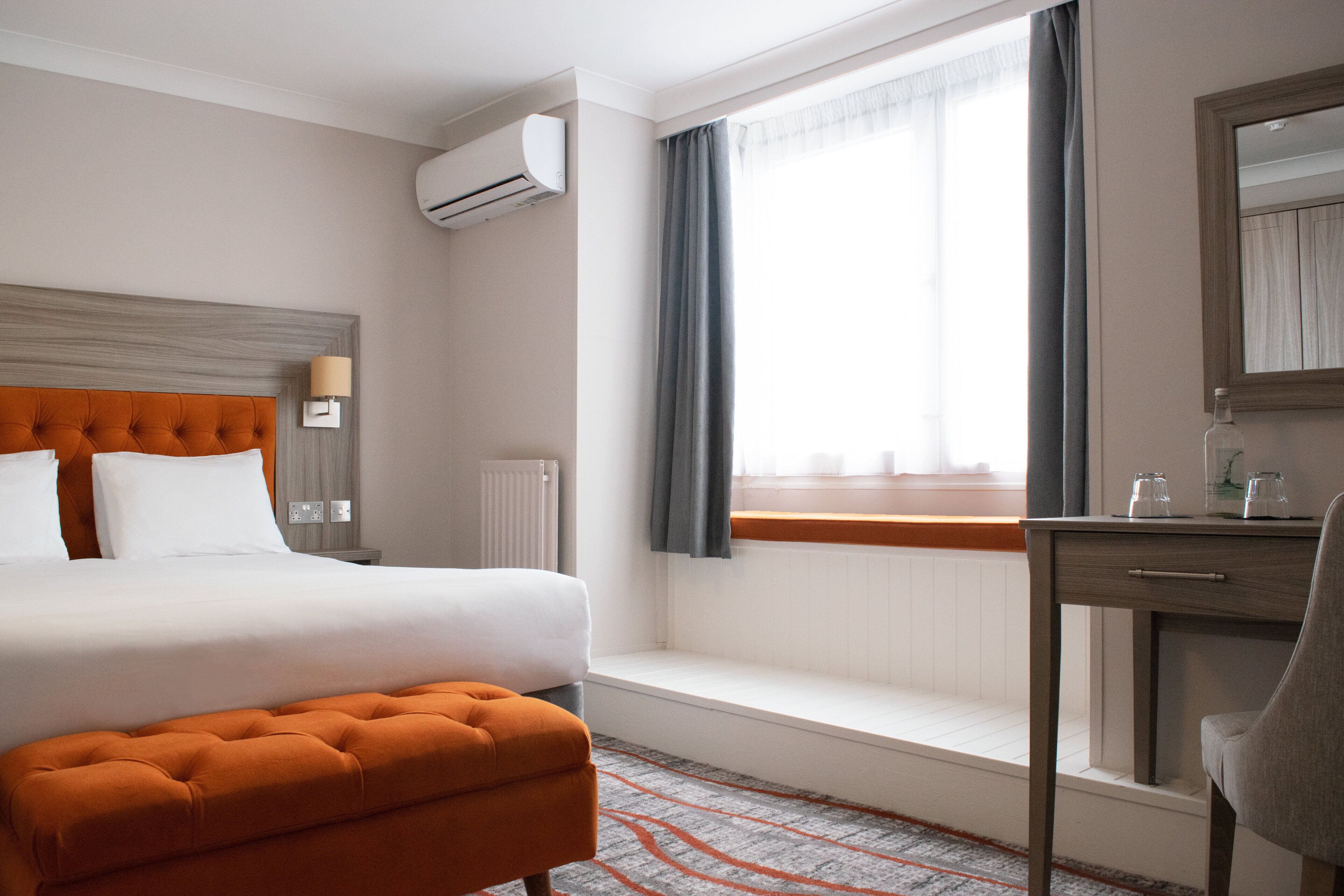 A hotel room with a bed, a desk, a chair, a window with curtains, an air conditioning unit, and a radiator.