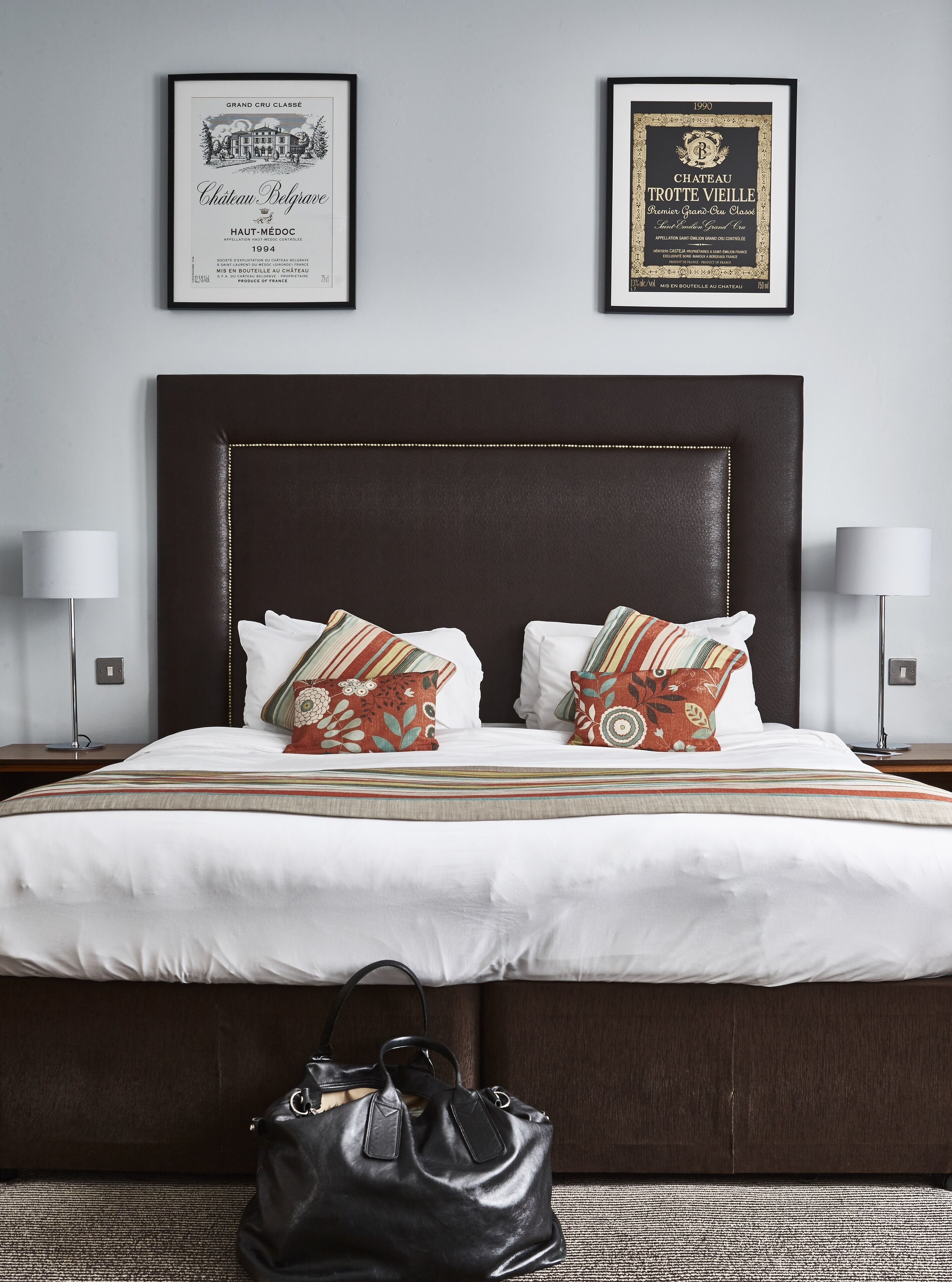 Een netjes opgemaakt bed met een donker hoofdbord, twee kussens en een gestreept dekbedovertrek. Boven het bed hangen twee gecertificeerde lijsten aan de muur.