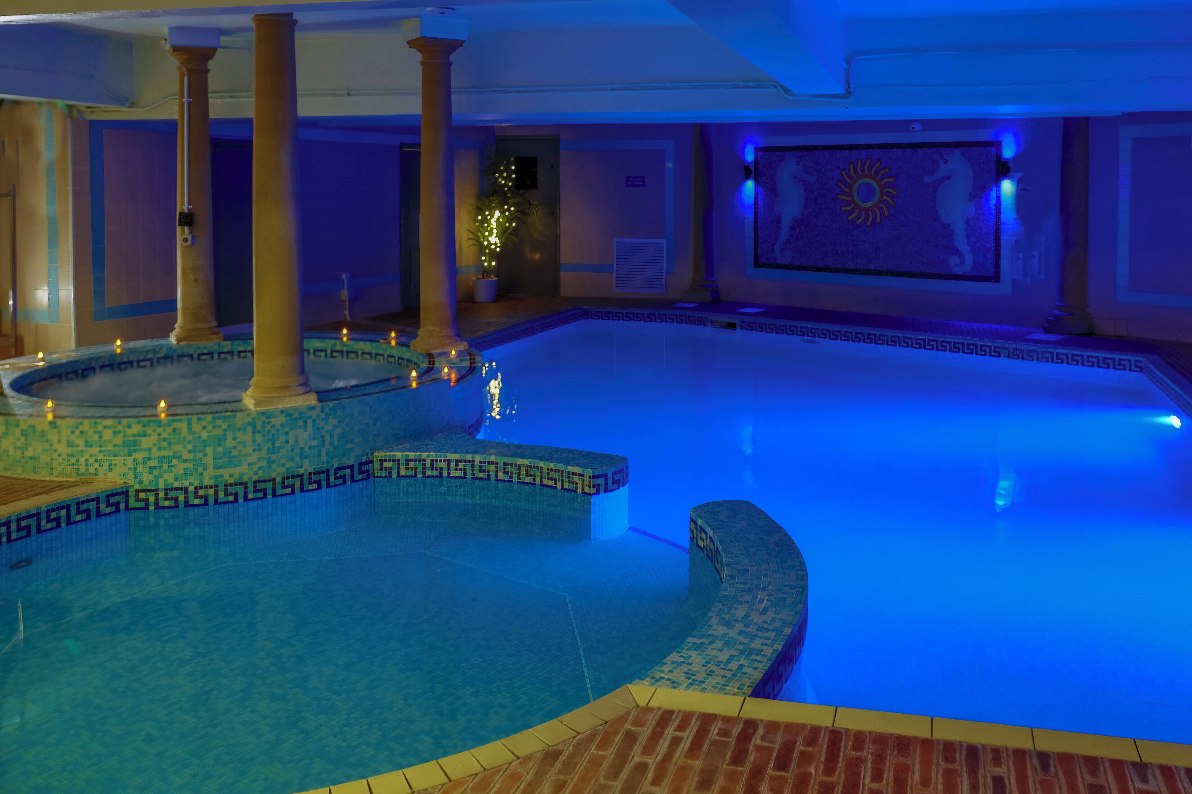 Ein großer, überdachter Swimmingpool mit blau beleuchtetem Design, inklusive eines Mosaikmusters an den Wänden und einem Wandgemälde mit Sonnenmotiv.