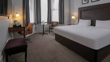 A hotel room with a large bed, a desk with a lamp, a chair, a small table with bottles, and a window with curtains.