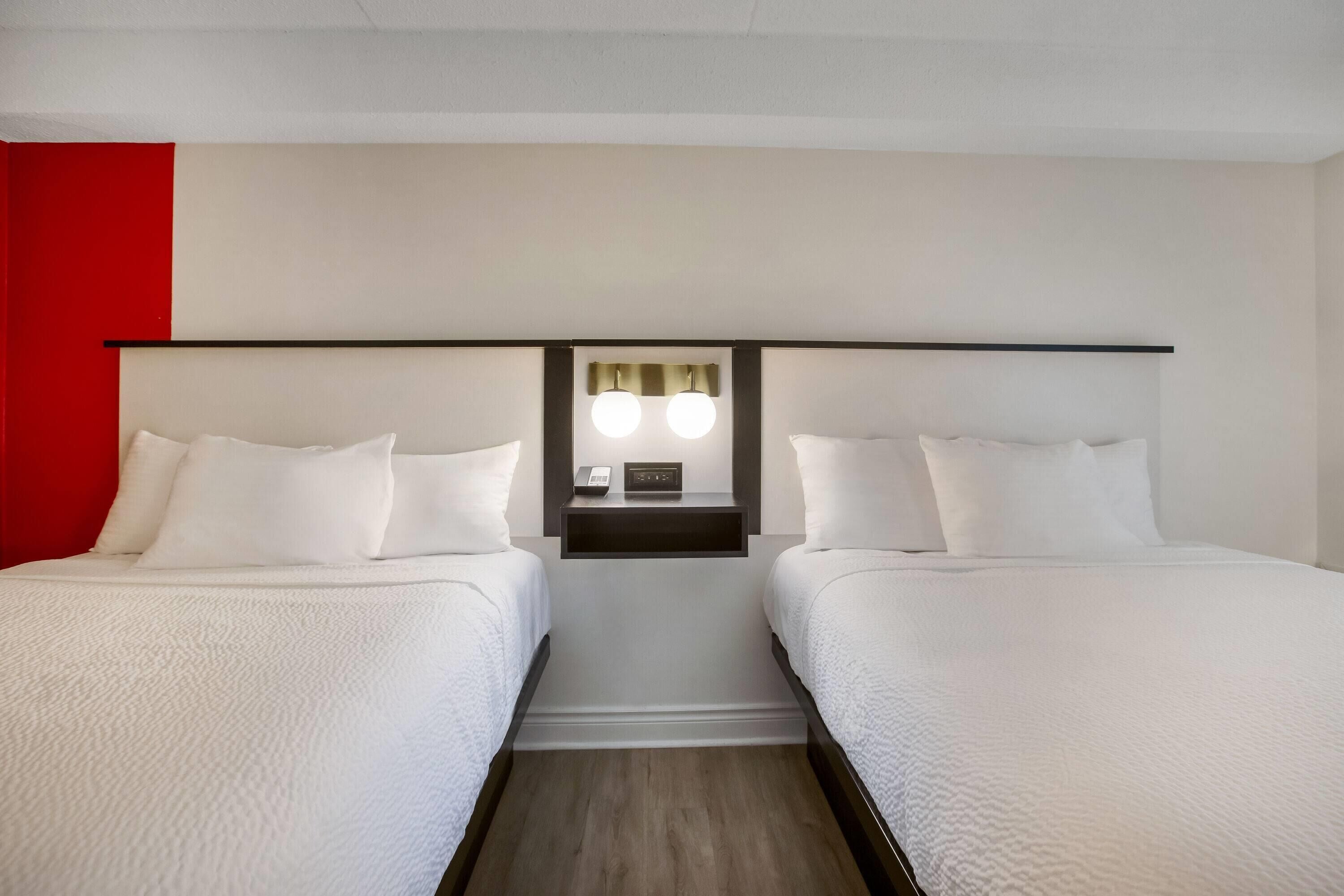 Quarto de hotel com duas camas, uma parede de destaque vermelha, uma mesinha de cabeceira com abajur e um criado-mudo com telefone.