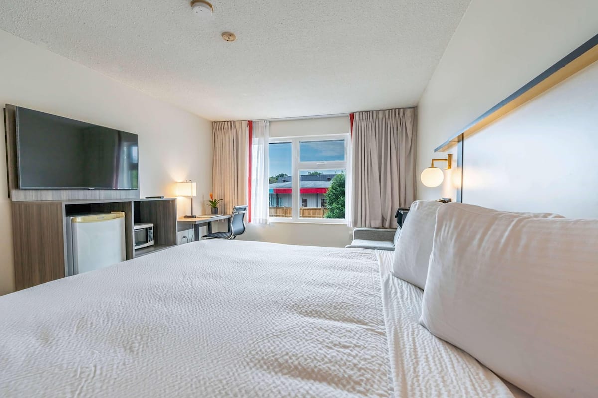 A hotel room with a bed, a TV, a desk with a chair, a window with curtains, and a view of the outside.