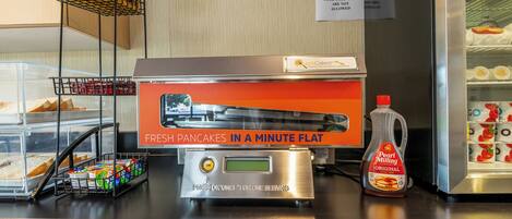 A breakfast counter with a pancake maker, a bottle of syrup, and a sign indicating fresh pancakes in a minute flat.