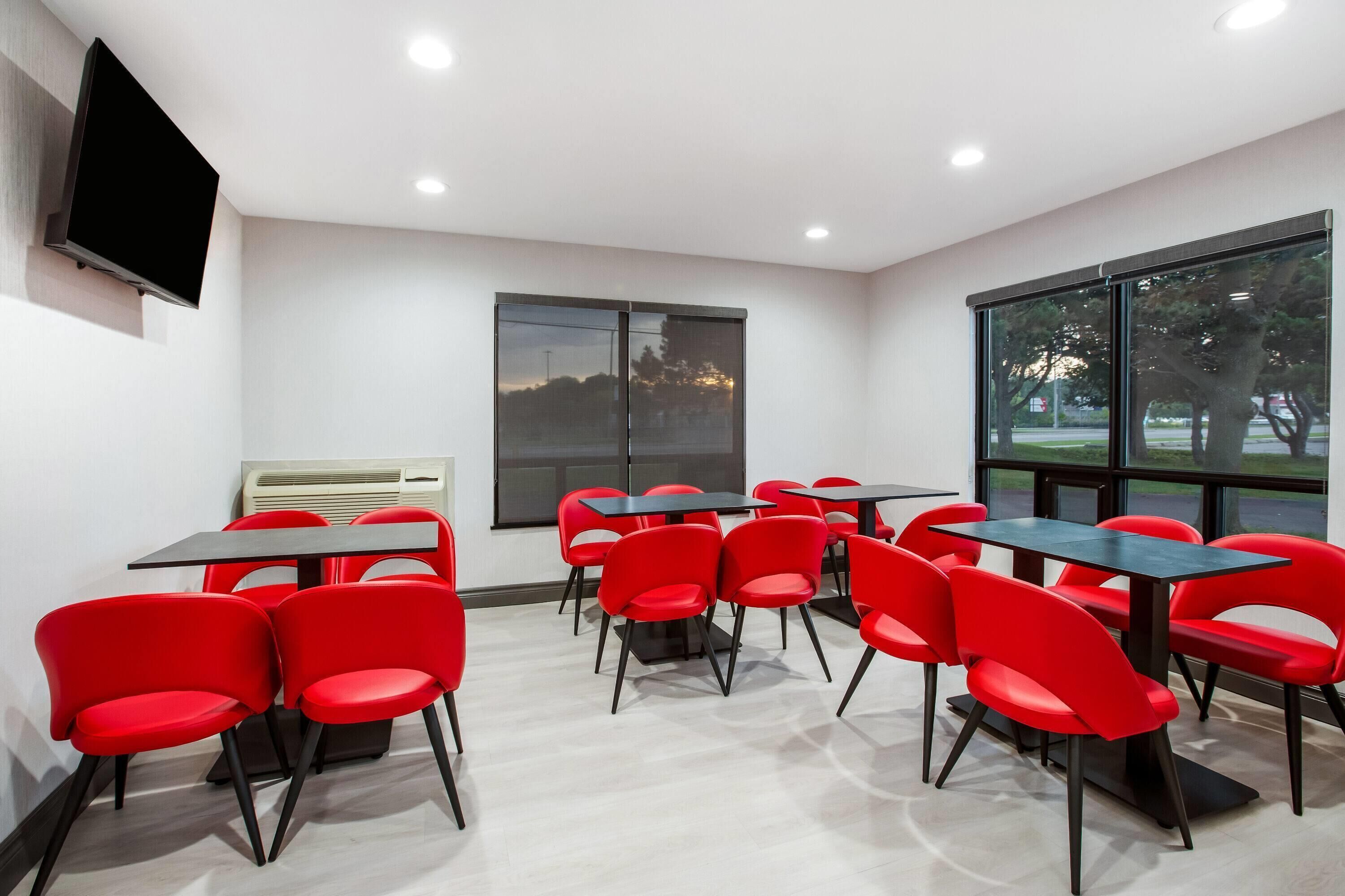 A conference room with red chairs and tables, a flat-screen TV, and a large window with a view of trees and a building.