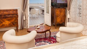 A hotel room with a bed, a TV, a wooden side table, a white armchair, a chandelier, and a view of the sea through an open door.