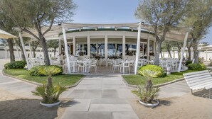 A covered outdoor seating area with white tables and chairs, surrounded by greenery and trees.