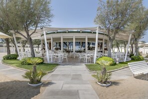 A covered outdoor seating area with white tables and chairs, surrounded by greenery and trees.