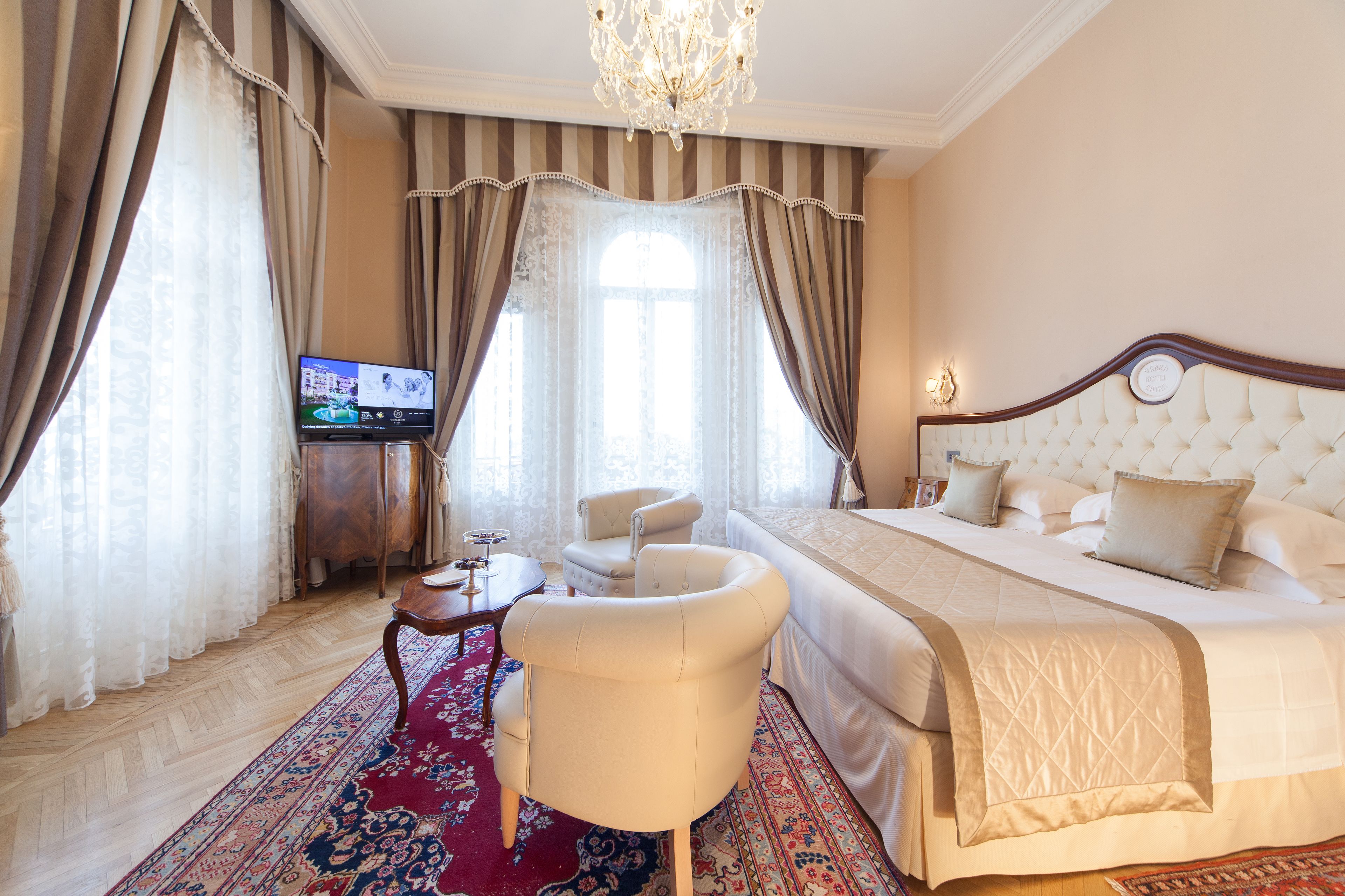 A hotel room with a large bed, a sitting area with a chair, a television, and a chandelier.