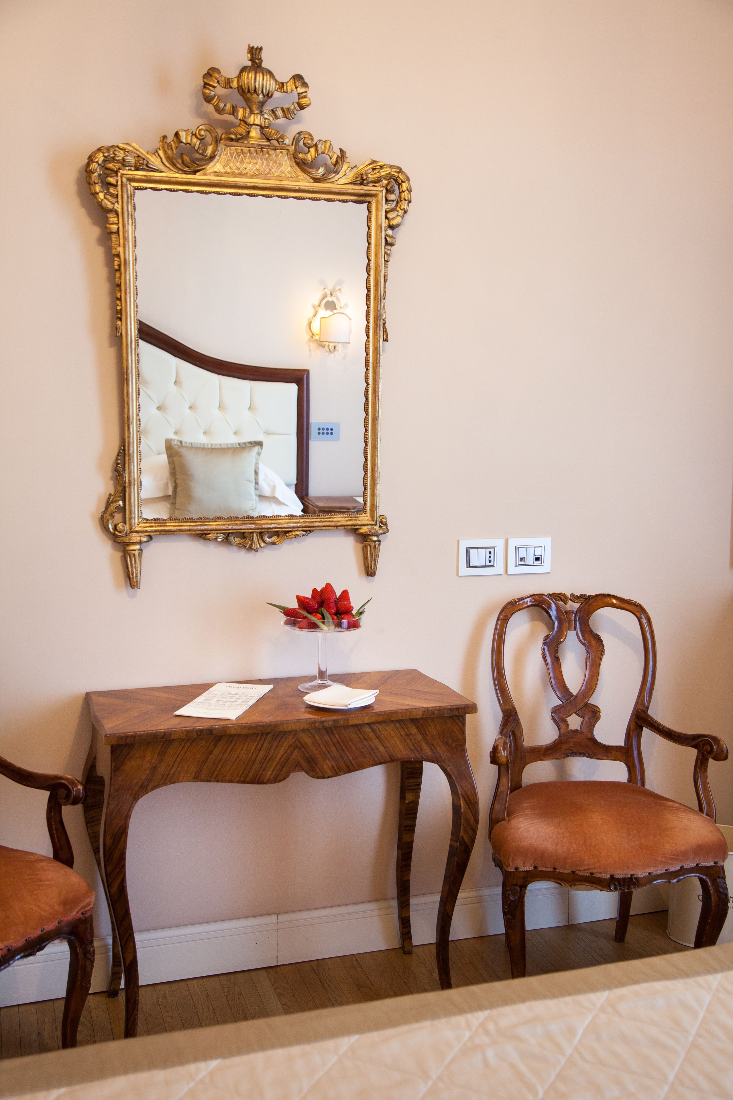 Una habitación con un espejo grande y ornamentado, una mesa de madera con un jarrón de flores rojas, dos sillas de madera y una cama con cubrecama acolchado.