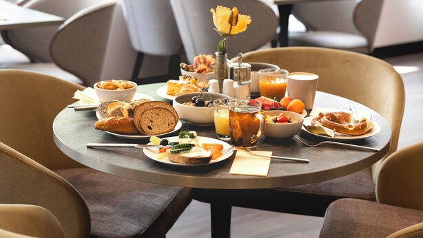 A breakfast setting with a variety of food items, including bread, fruits, and a beverage, on a round table in a modern dining area with chairs. - Leonardo Hotel Heidelberg City Center (Heidelberg)