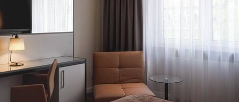 Una habitación de hotel con una cama, un escritorio con silla, televisión, una mesita y una ventana con cortinas.