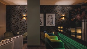 A modern lounge area with patterned wallpaper, a bar with a view, and a seating area with green and blue chairs. - Leonardo Hotel Heidelberg City Center (Heidelberg)