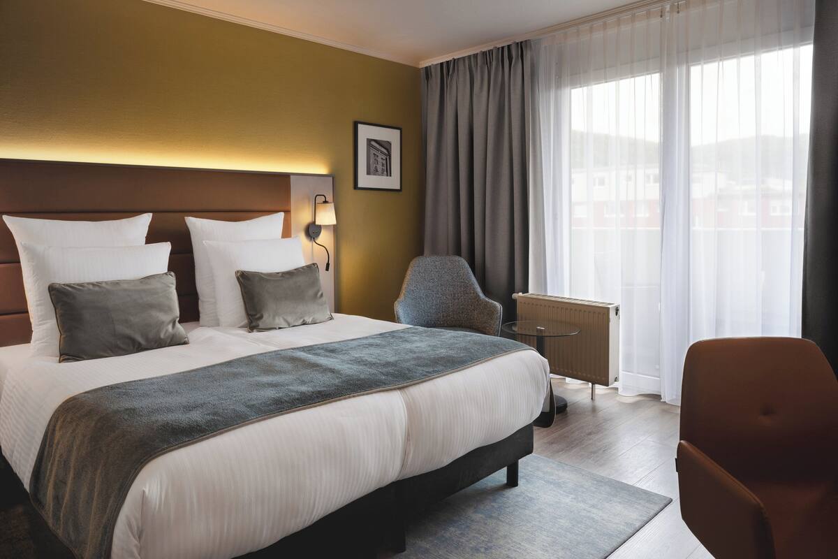 Une chambre d’hôtel avec un grand lit, une chaise, un radiateur et une fenêtre avec des rideaux.