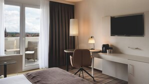 Habitación de hotel con cama, escritorio con lámpara, una silla, televisor y vista a la ciudad a través de amplias ventanas.