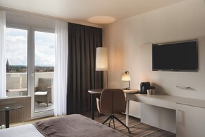 Hotelkamer met een bed, een bureau met lamp, een stoel, een televisie en uitzicht op de stad door grote ramen.