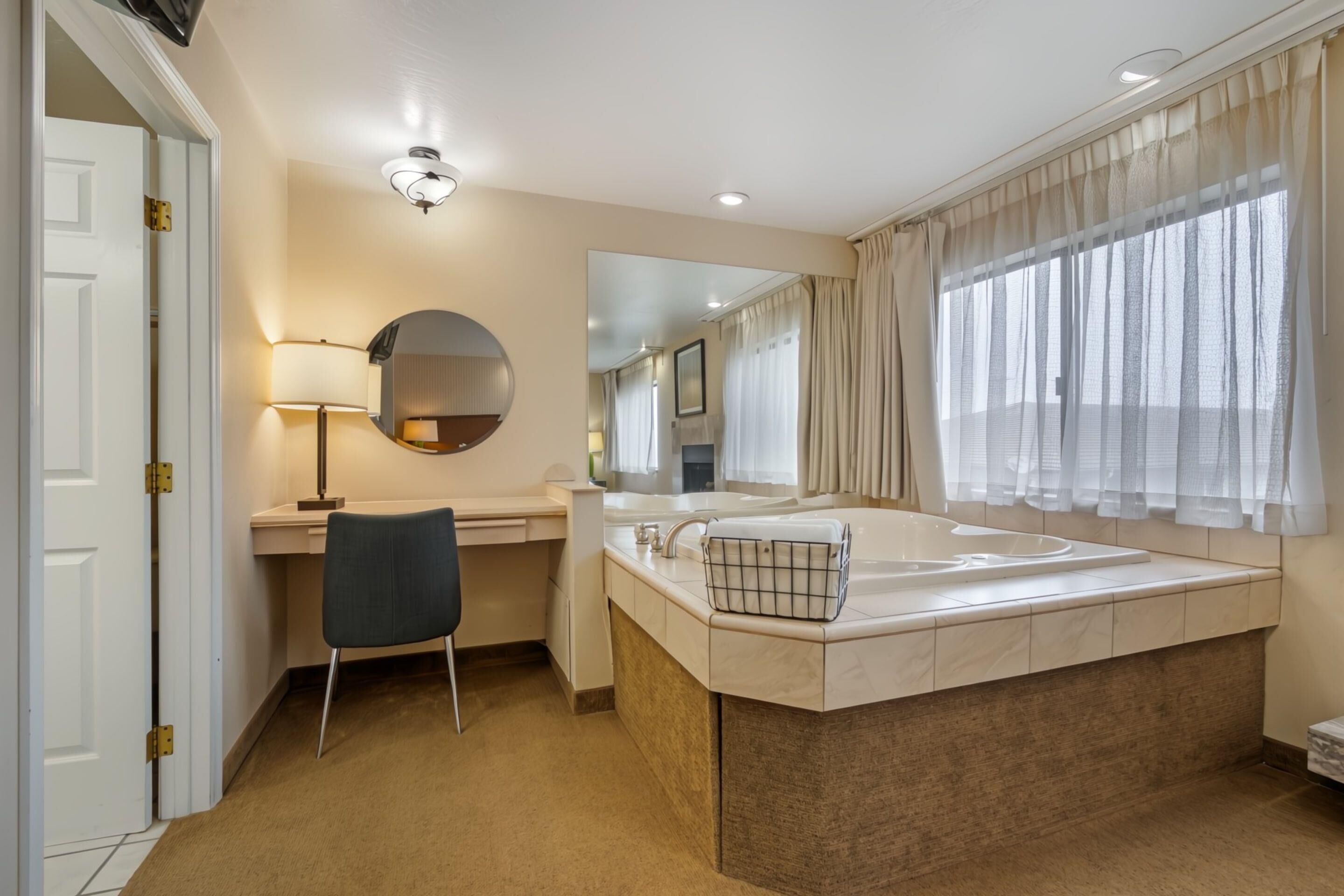 A modern bathroom with a large bathtub, a round mirror, a desk with a chair, and a window with sheer curtains.