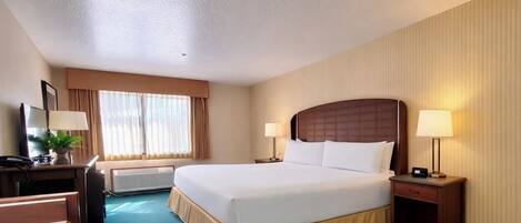 A hotel room with a large bed, two bedside tables with lamps, a desk with a chair, a radiator, and a window with curtains.