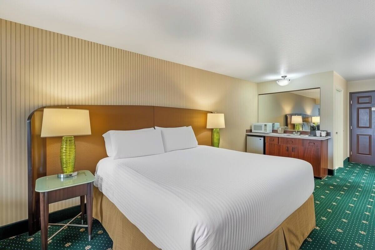 Ein Hotelzimmer mit einem großen Bett, Nachttischlampen, einem kleinen Tisch, einem Spiegel, einer Küchenzeile und einer Tür.