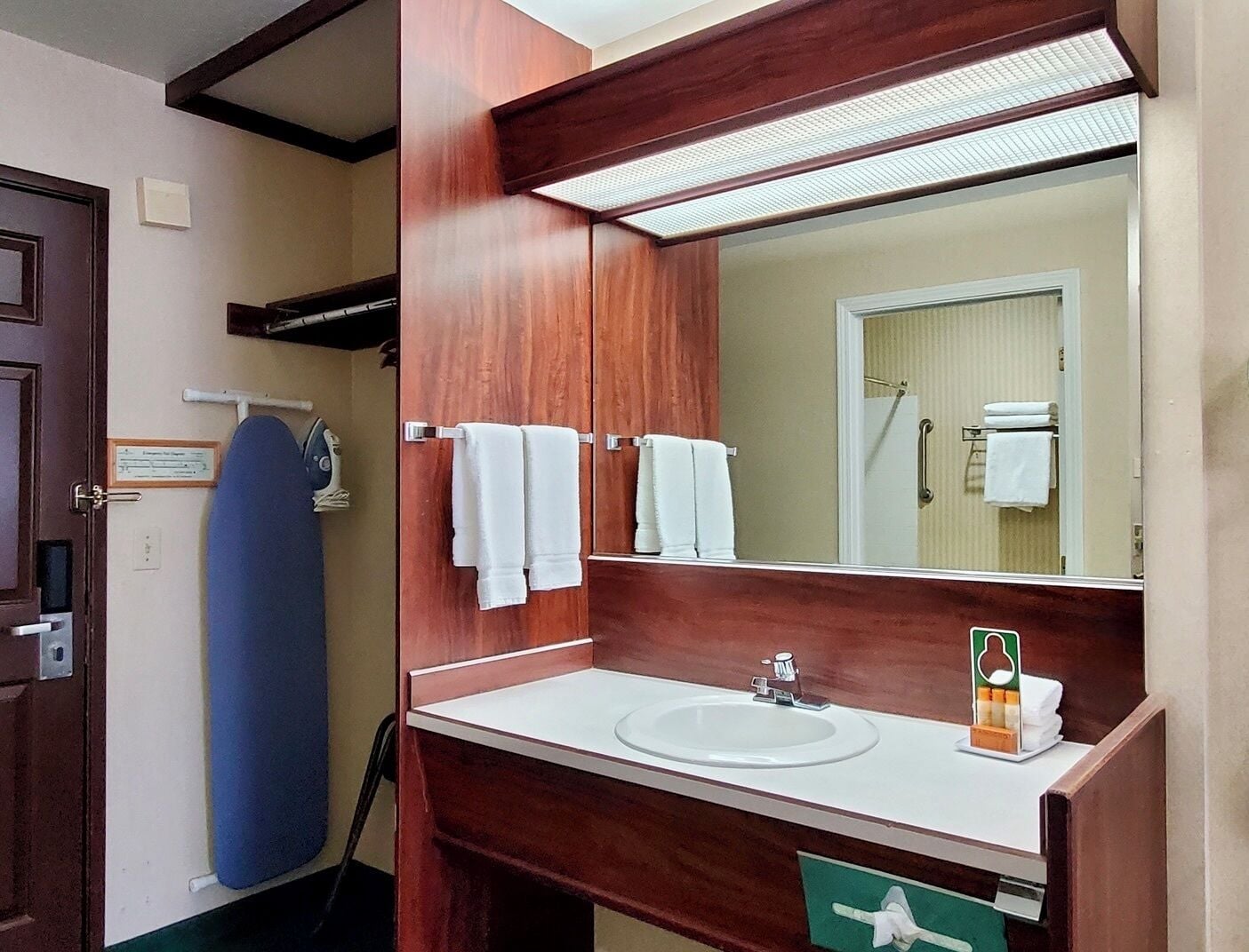 A bathroom with a wooden vanity, a white sink, and a mirror. There is a towel rack with white towels, a blue surfboard, and a green trash bin.