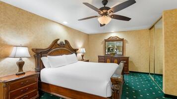 Habitación de hotel con una cama de madera, dos mesitas de noche con lámparas, un ventilador de techo, un espejo grande y una alfombra verde estampada.