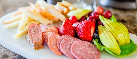 A charcuterie board with sliced meats, fruits, and cheese, accompanied by two bottles and a glass of wine on a marble table.