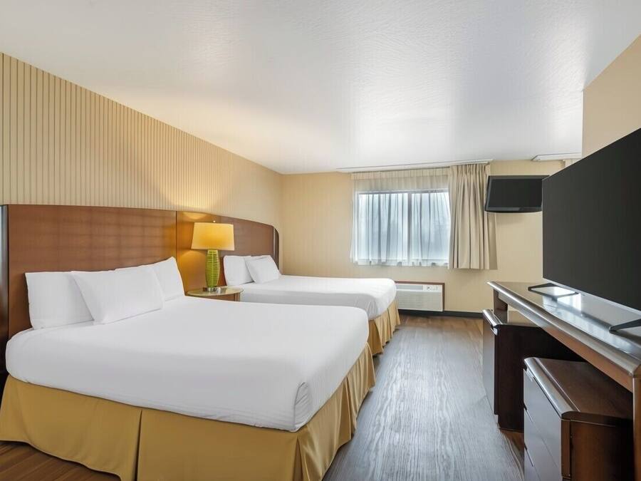 A hotel room with two beds, a desk with a flat-screen TV, a lamp, and a window with curtains.