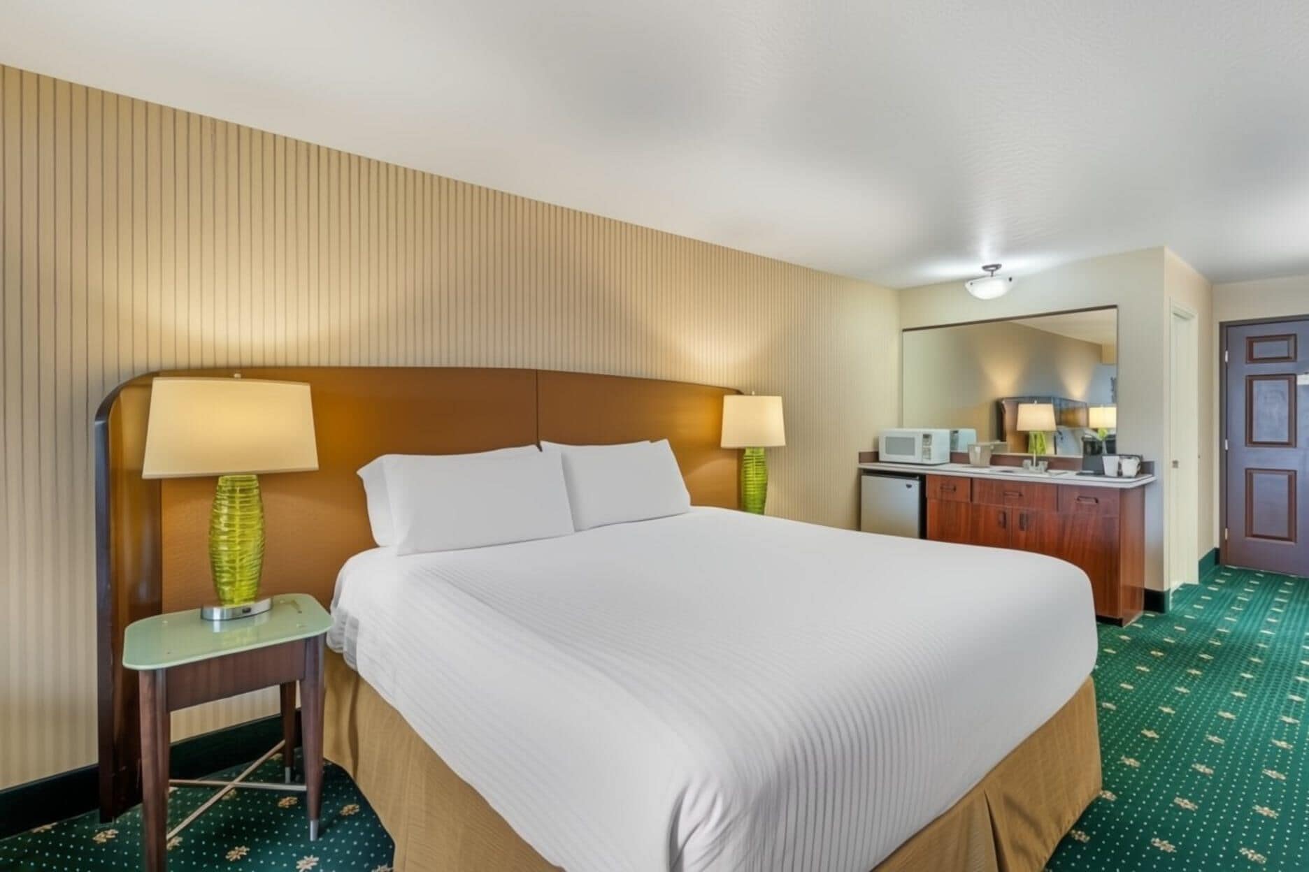 Quarto de hotel com cama grande, duas luminárias de cabeceira, uma mesinha, um espelho, micro-ondas e guarda-roupa.
