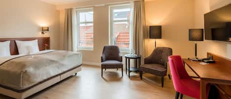A hotel room with a bed, two armchairs, a small table, a desk with a chair, a TV, and two windows with curtains.