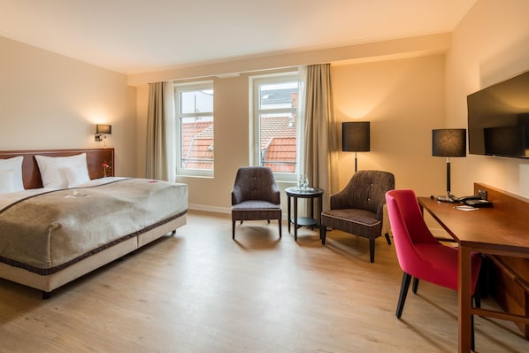 Een hotelkamer met een bed, twee fauteuils, een kleine tafel, een bureau met stoel, een televisie en twee ramen met gordijnen.