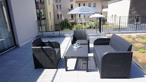 An outdoor seating area with wicker furniture and a table, a striped umbrella, and a view of residential buildings in the background. - Best Western Plus Hotel Excelsior (Erfurt)