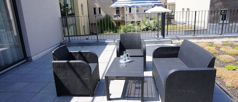 An outdoor seating area with wicker furniture and a table, a striped umbrella, and a view of residential buildings in the background.