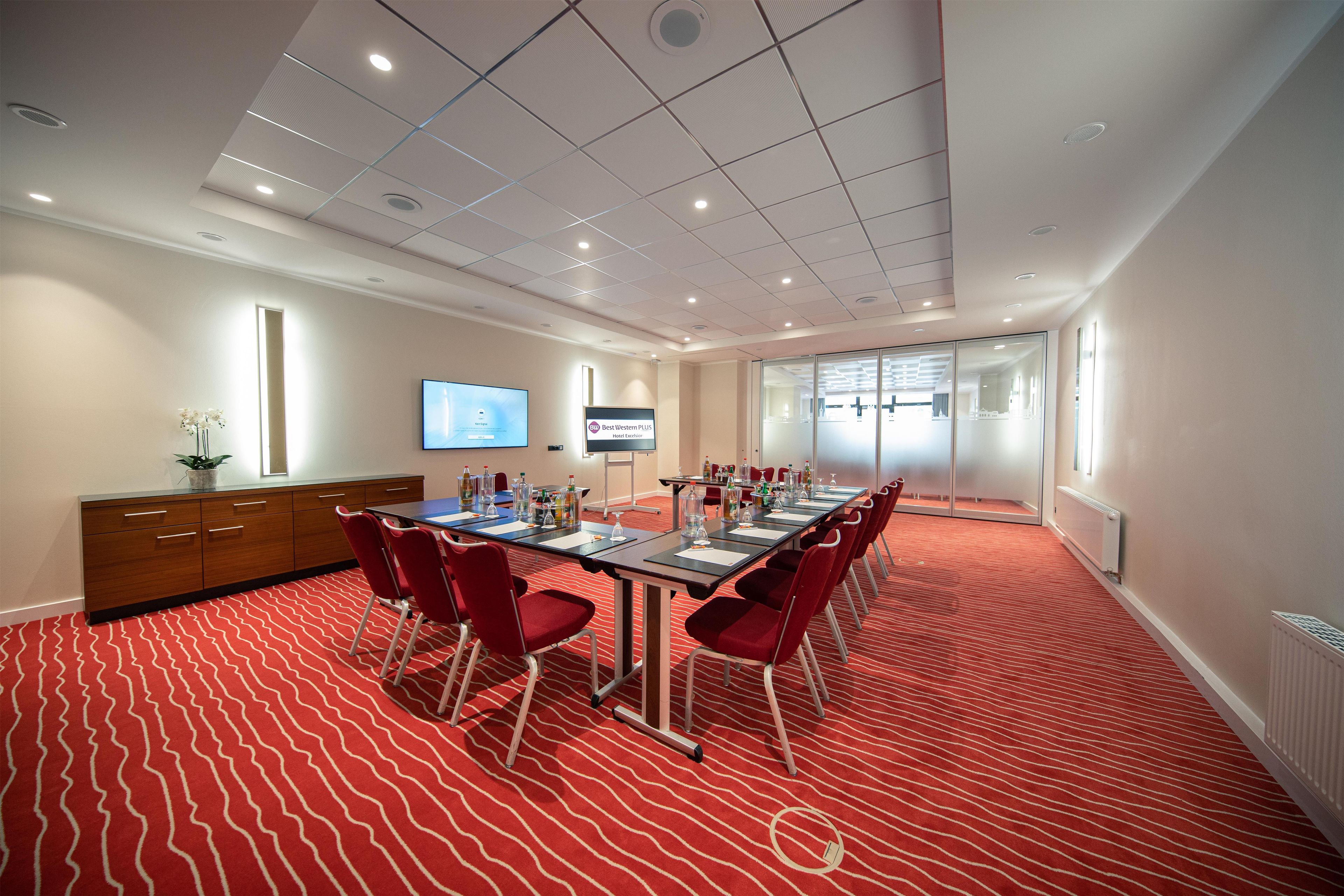 Un salón de conferencias con una mesa larga, sillas rojas, una pantalla de televisión y una pantalla digital en la pared.