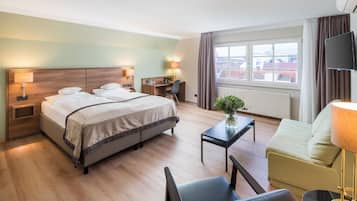 Una habitación de hotel con cama, mesitas de noche, escritorio, silla, sofá, televisión y ventana con cortinas.