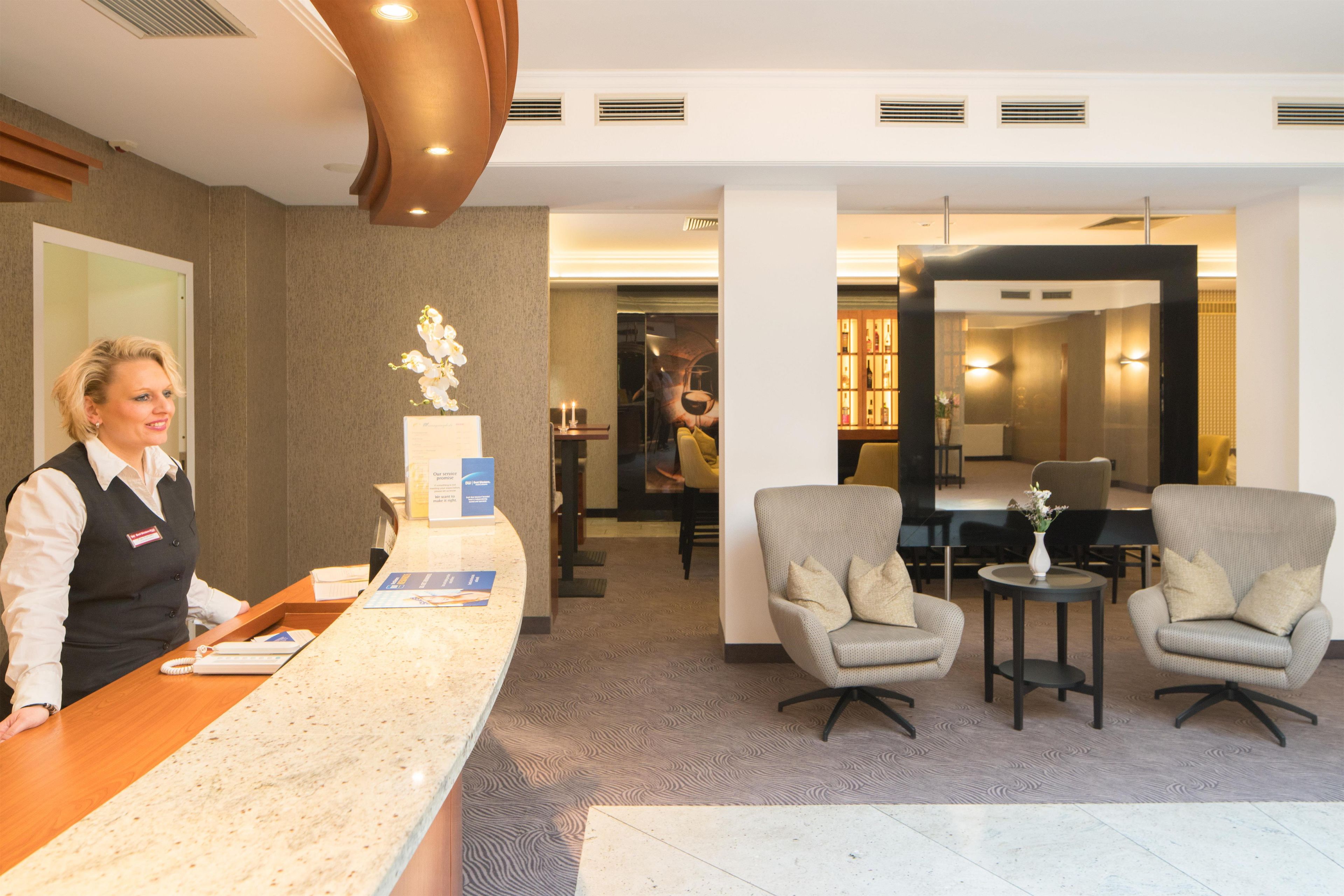 Zona de recepción de un hotel con una recepcionista, mostrador de recepción, dos sillones, una mesita y un espejo.